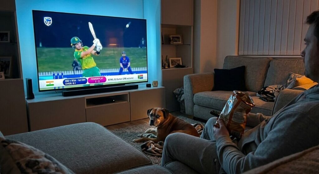 A man watches cricket on a large TV while his dog gives him a judgmental side-eye from the floor.