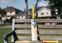 Cricket Gear List: What Every Cricket Player Should Own colorful cricket bat leaning against the slats, white batting pads draped over the armrest, a dark blue helmet resting on the seat, and a red cricket ball nearby, with suburban houses, trees, and a fence blurred softly in the background.